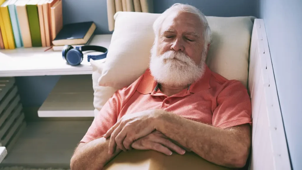 A senior man with a white beard rests his head back on a pillow with closed eyes to recover from a daytime energy crash.