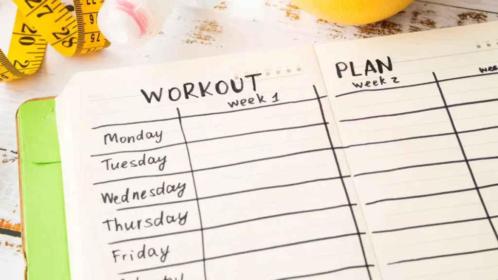 Open workout journal showing a weekly fitness schedule, paired with a water bottle and measuring tape on a white wood desk.