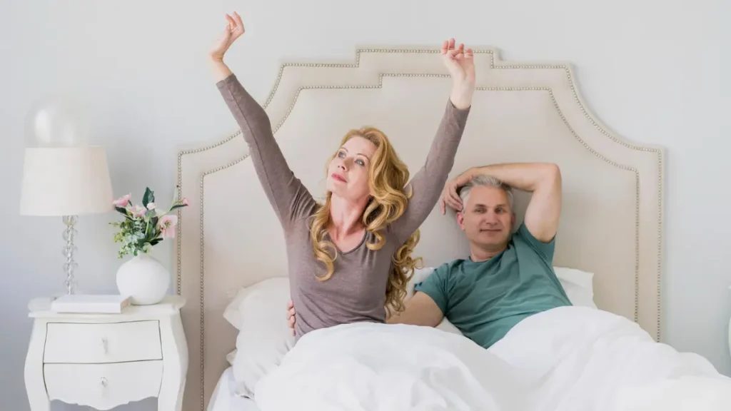 Couple in their 50s waking up refreshed, woman stretching arms up, man smiling, showing better sleep after fixing caffeine habits.