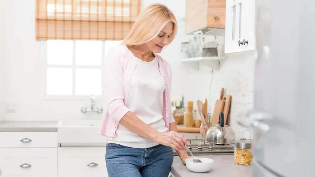 Blonde woman in her 50s stirring psyllium husk fiber into a bowl at a bright kitchen counter as part of a morning gut health routine.