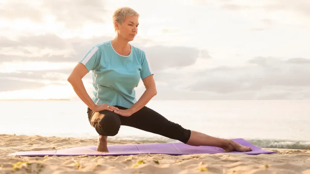 Older woman holding a low lunge yoga pose on a mat at the beach, focused and steady in soft morning light.