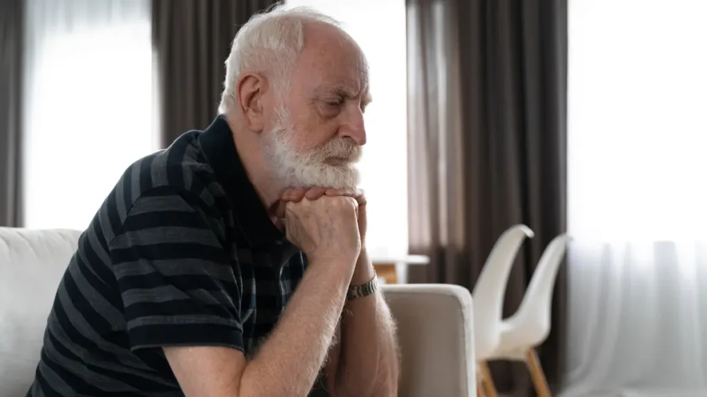 An elderly man sits resting his chin on his hands, looking exhausted and restless while trying to wind down in a living room.