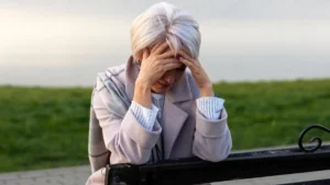 An older woman with short white hair sits on a park bench, head in her hands, expressing exhaustion or distress, wearing a gray coat and plaid scarf.