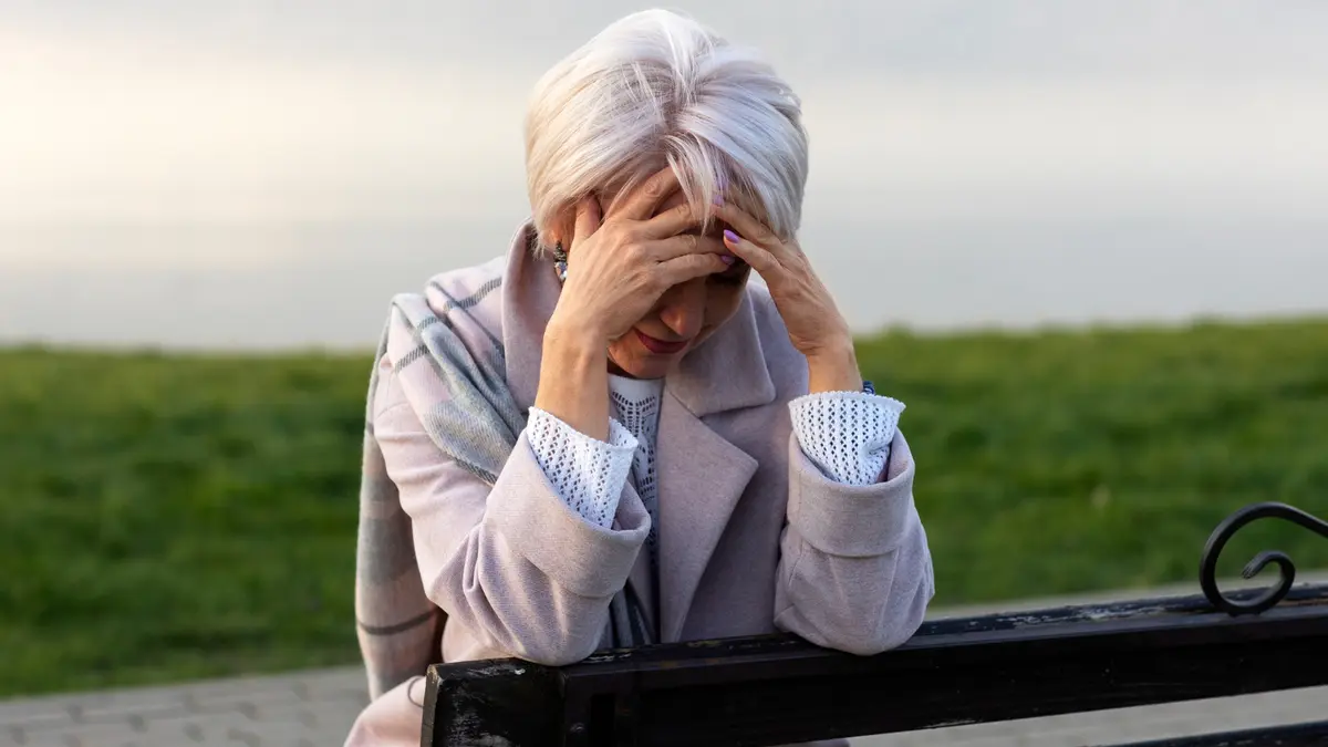 An older woman with short white hair sits on a park bench, head in her hands, expressing exhaustion or distress, wearing a gray coat and plaid scarf.