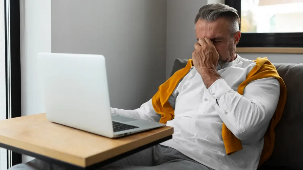 A fatigued man over 50 sitting at a laptop and rubbing his eyes, illustrating brain fog and exhaustion from nutrient deficiency.