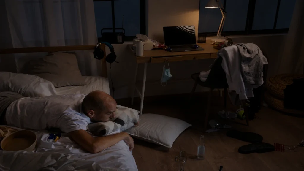 Man sleeping in a cluttered bedroom with laptop and work items, how bedroom clutter disrupts deep sleep after 50.