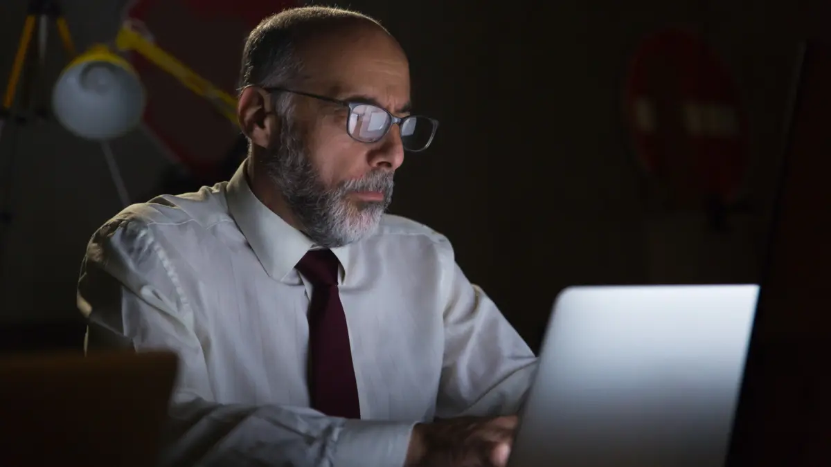 A middle-aged man wearing glasses works on a laptop in a dimly lit room, with screen light illuminating his face.