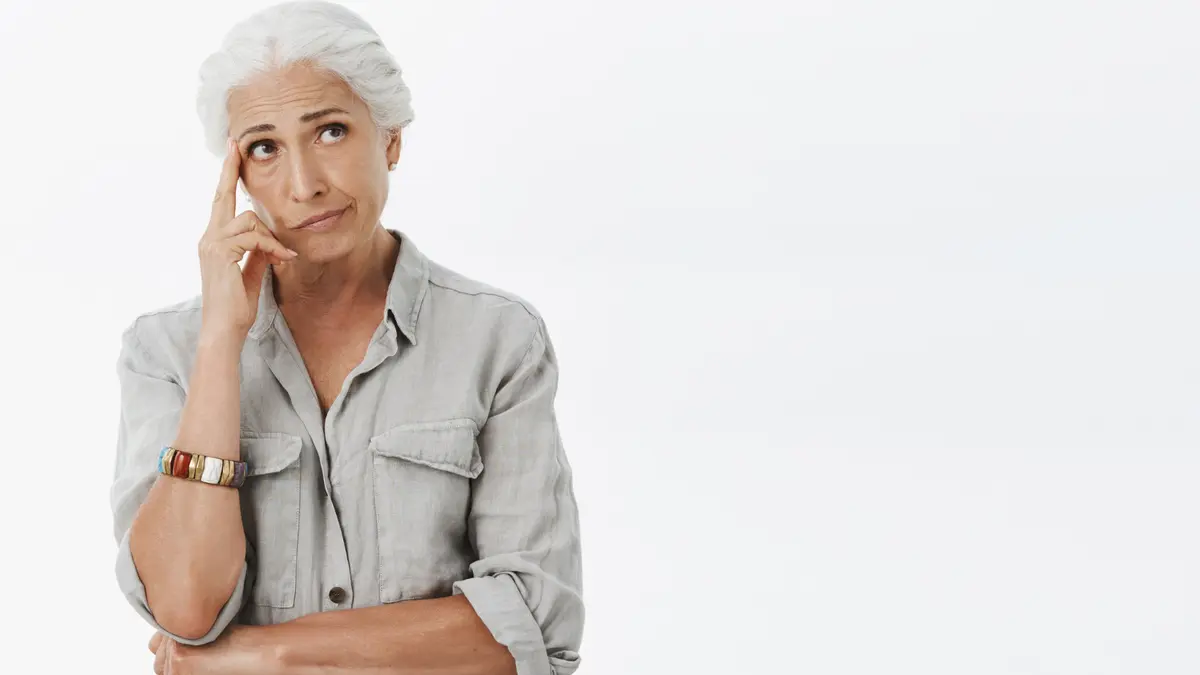 Older woman with white hair thinking thoughtfully, finger to temple, weighing her options against a clean background.
