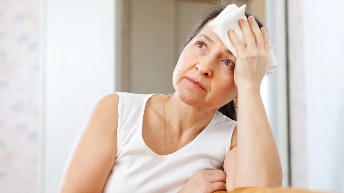 A mature woman with grey hair presses a folded white cloth to her forehead with a weary, pensive expression, visually representing the hidden fatigue often caused by hypothyroidism after 50.