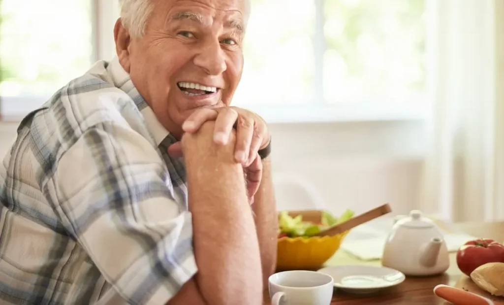 Older man smiling at a breakfast table with a small cup of coffee, fresh vegetables, and a teapot, representing healthy caffeine habits after 50.