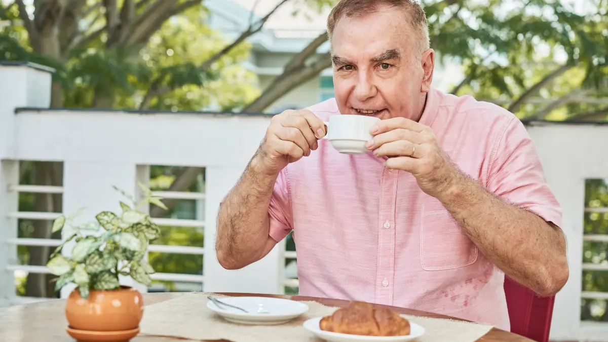 A cheerful older man in a pink shirt sips from a small coffee cup at an outdoor table with a croissant beside him illustrating how enjoyable afternoon caffeine habits can quietly disrupt sleep hours later.