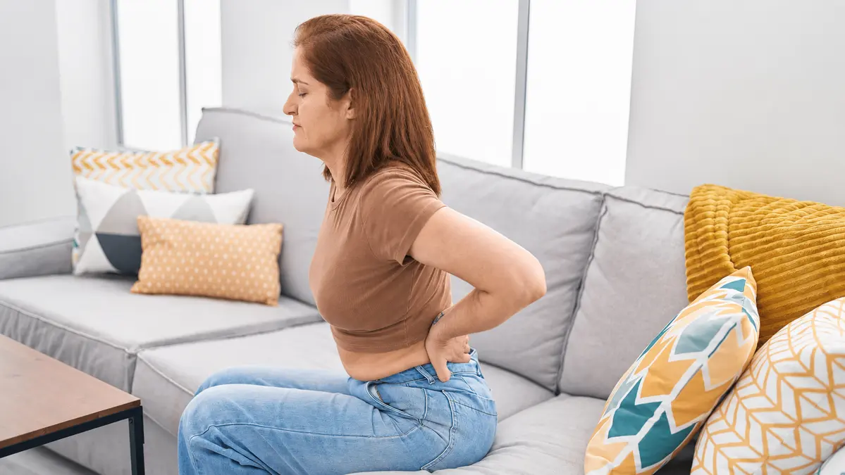 A woman over 50 sits on a couch gripping her lower back, showing kidney discomfort that water alone cannot prevent.