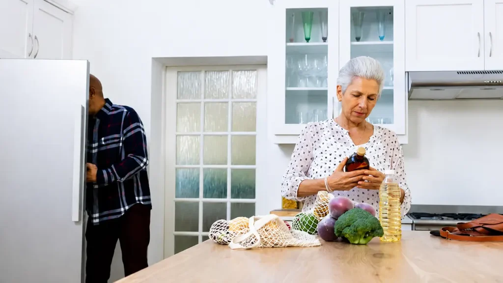 Silver-haired woman in her 60s reading the ingredient label on a bottle at a kitchen counter, with fresh onions and broccoli nearby, checking for fast-fermenting fiber sources.