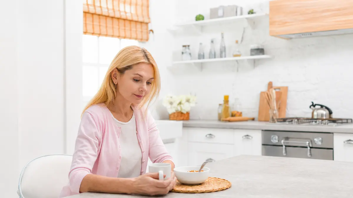 Woman over 50 sitting at kitchen table with coffee and breakfast bowl, looking down with a quietly frustrated expression despite eating well.