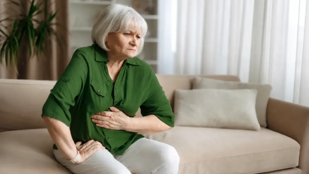 Woman in her early 60s on a couch, hand on abdomen, expression showing quiet concern while recognizing gut dysbiosis symptoms after antibiotics.