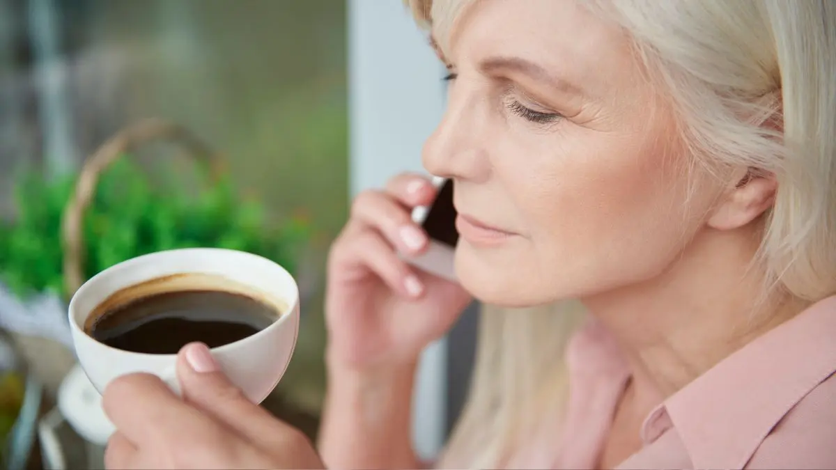 Woman in her 60s holding a white cup of black coffee while on a phone call — daily coffee consumption shown to reduce uric acid levels by blocking xanthine oxidase, the enzyme that converts purines into uric acid.