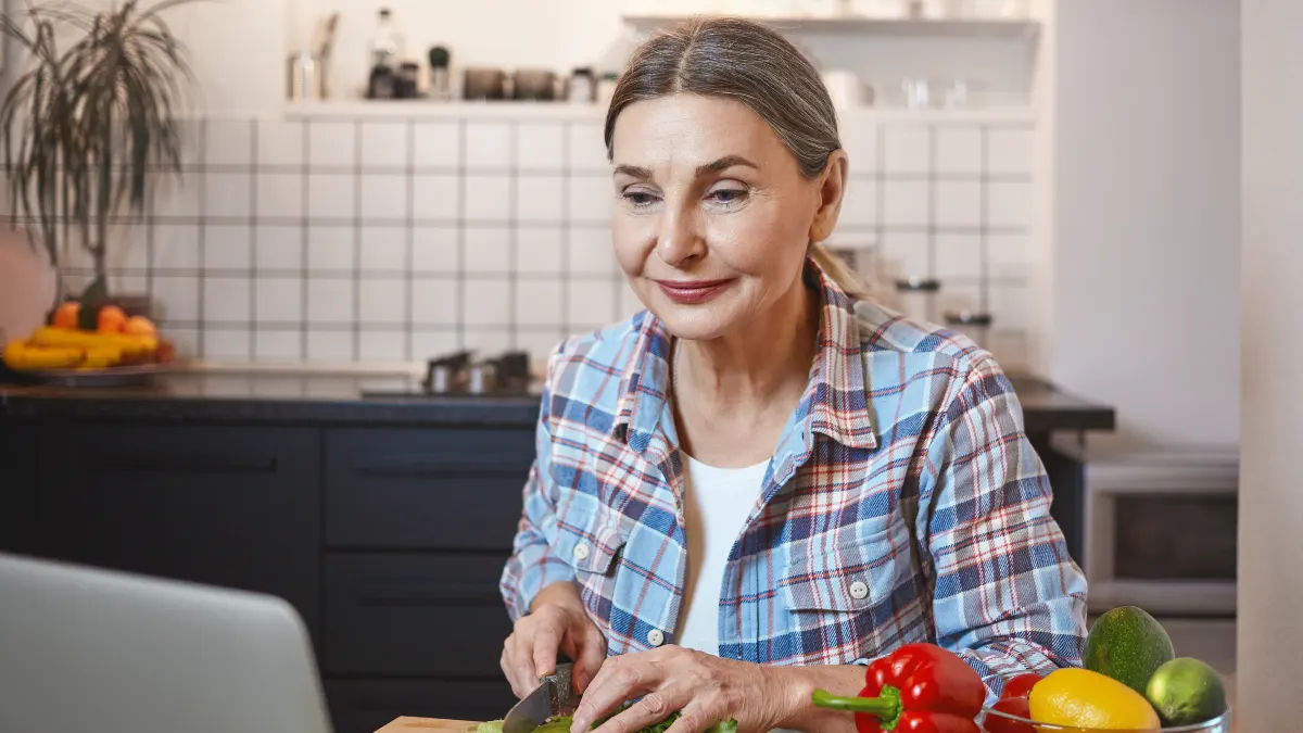Mature woman in a flannel shirt using a laptop to follow a new recipe while chopping fresh vegetables in a bright kitchen.