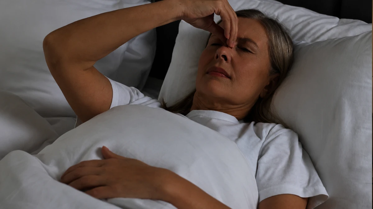 Mature woman lying awake in a dark bedroom at 2 AM, pressing her hand to her face with the quiet frustration of middle-of-the-night waking after 50.