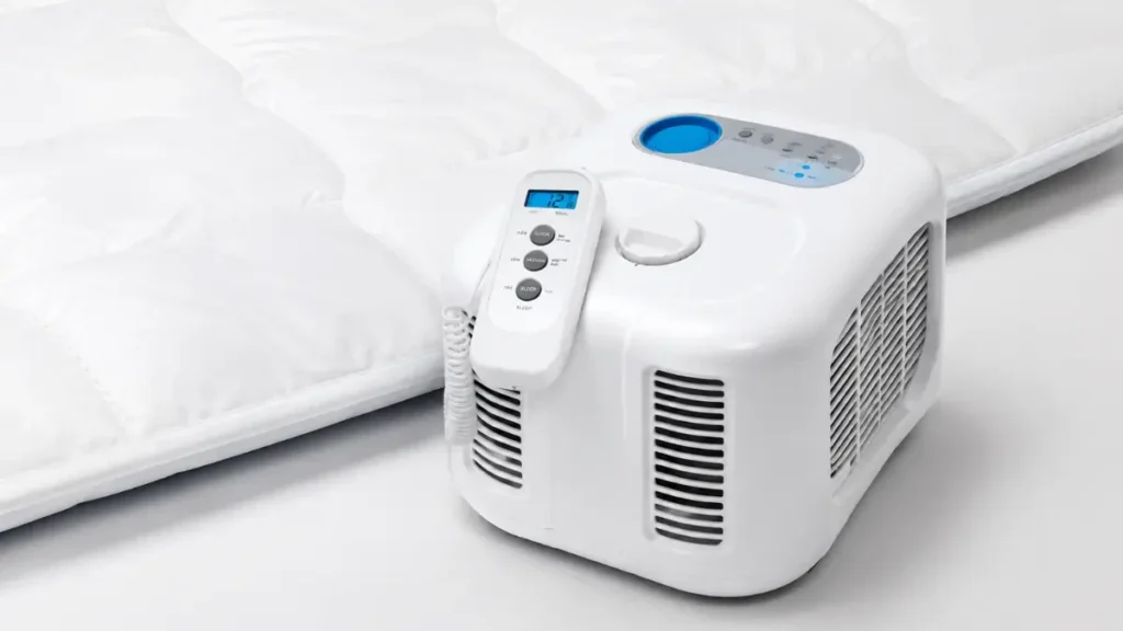 A water-cooled mattress pad control unit with a remote beside a white mattress pad, representing cooling sleep systems that target body temperature directly without cooling the whole room.