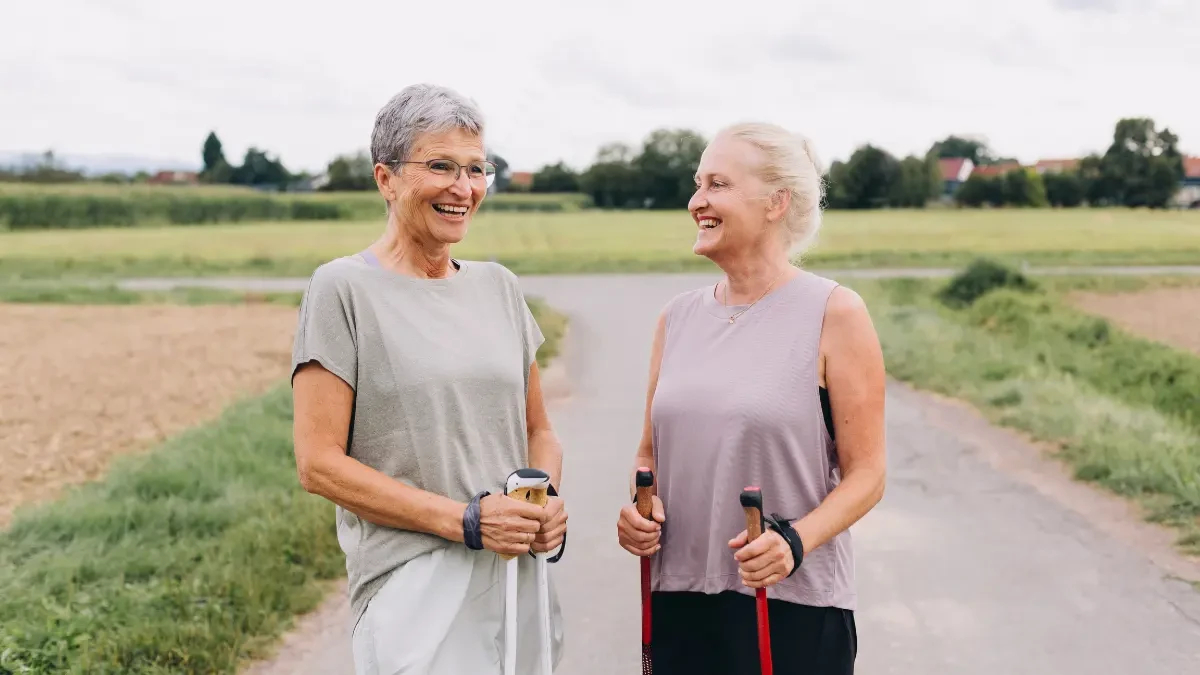 Two women in their late 50s laughing and walking together on a sunny outdoor path, showing how daily steps support healthy aging after 50.