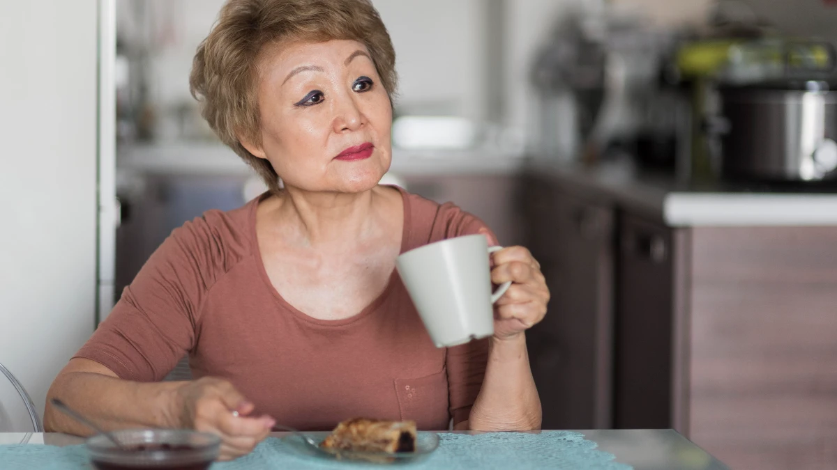 Thoughtful mature Asian woman sitting alone at a kitchen table with a simple meal and coffee, illustrating the link between solo dining and heart health stress.