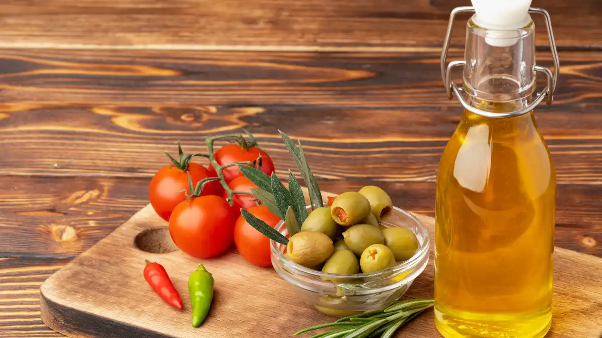A swing-top glass bottle of golden extra virgin olive oil next to a bowl of green olives, cherry tomatoes, and fresh rosemary on a wooden board. A visual representation of healthy Mediterranean diet staples.