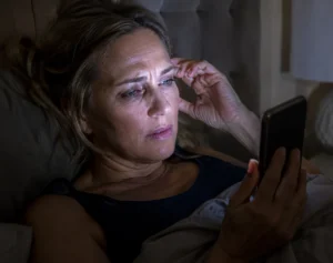 Woman in her 50s lying in bed at night rubbing her eye while looking at her phone screen in the dark, showing symptoms of digital eye strain and nighttime screen use discomfort.