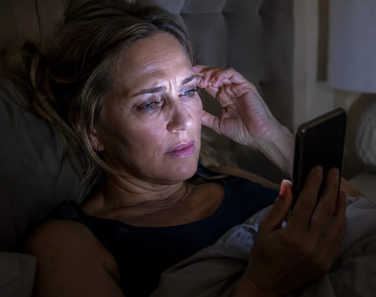 Woman in her 50s lying in bed at night rubbing her eye while looking at her phone screen in the dark, showing symptoms of digital eye strain and nighttime screen use discomfort.