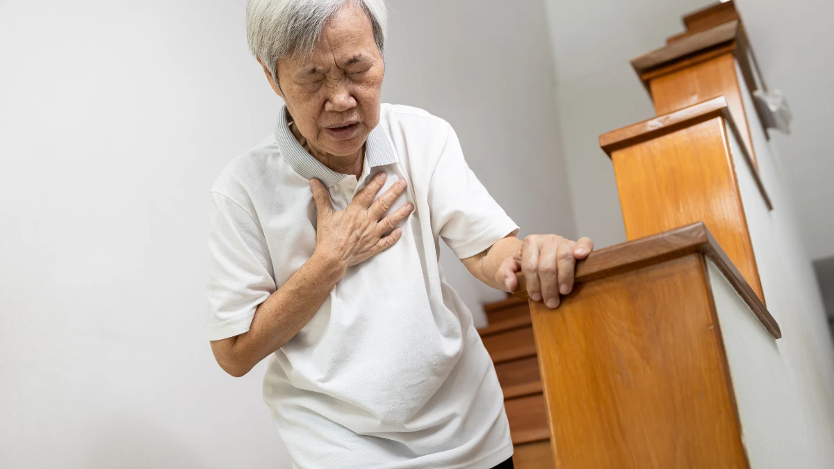 An older adult gripping a stair railing with one hand and pressing the other hand to their chest, showing signs of breathlessness and discomfort while climbing stairs.