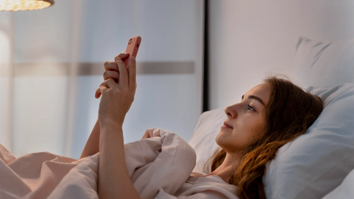 Woman lying in bed holding smartphone at arm's length in warm bedroom light, demonstrating proper phone distance to reduce eye strain.