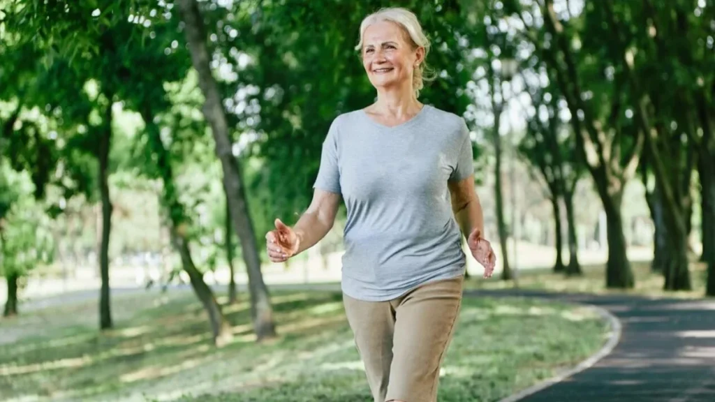 Woman in her early 60s walking confidently along an outdoor park path with arms swinging freely at her sides, demonstrating the improved balance and mobility that comes from consistent fall prevention training.