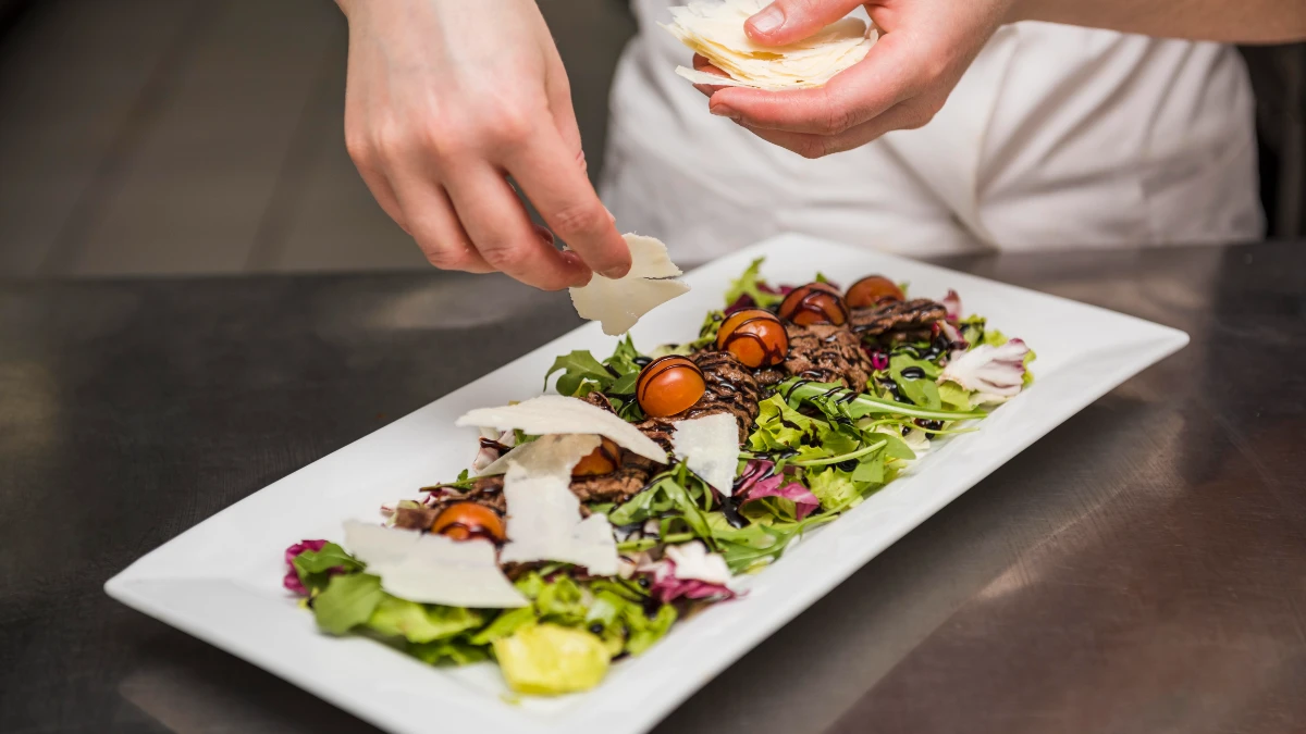 A chef places shaved hard cheese over a plated steak and mixed green salad to bind dietary oxalates in the stomach before digestion.