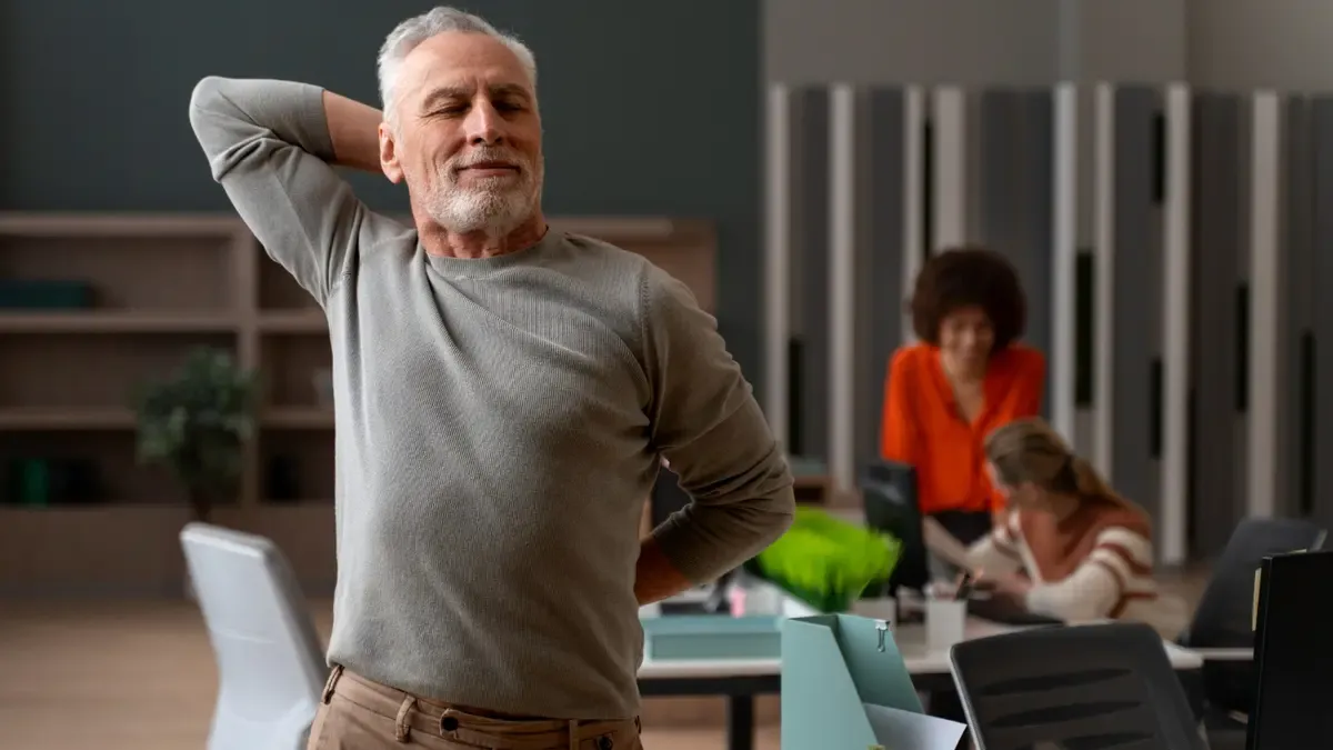 Older man standing at his office desk doing a neck stretch during a movement break, with colleagues working in the background.