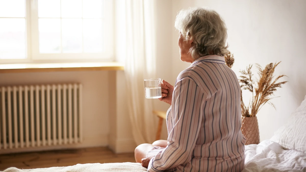An elderly woman in striped pajamas sits on the edge of a bed, viewed from behind, holding a glass of water while looking toward a sunlit window — illustrating the struggle of waking without a consistent morning routine.