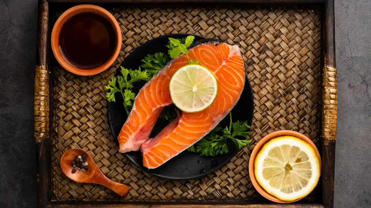 Fresh raw salmon steak with lemon and herbs on a woven tray, highlighting high-quality dietary sources of Omega-3 for muscle health.