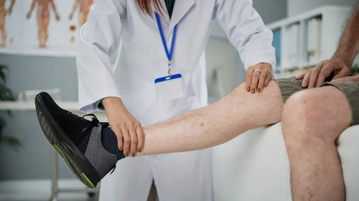 A doctor in a white coat examines a seated patient's leg and foot during a clinical assessment for nerve or circulation issues.