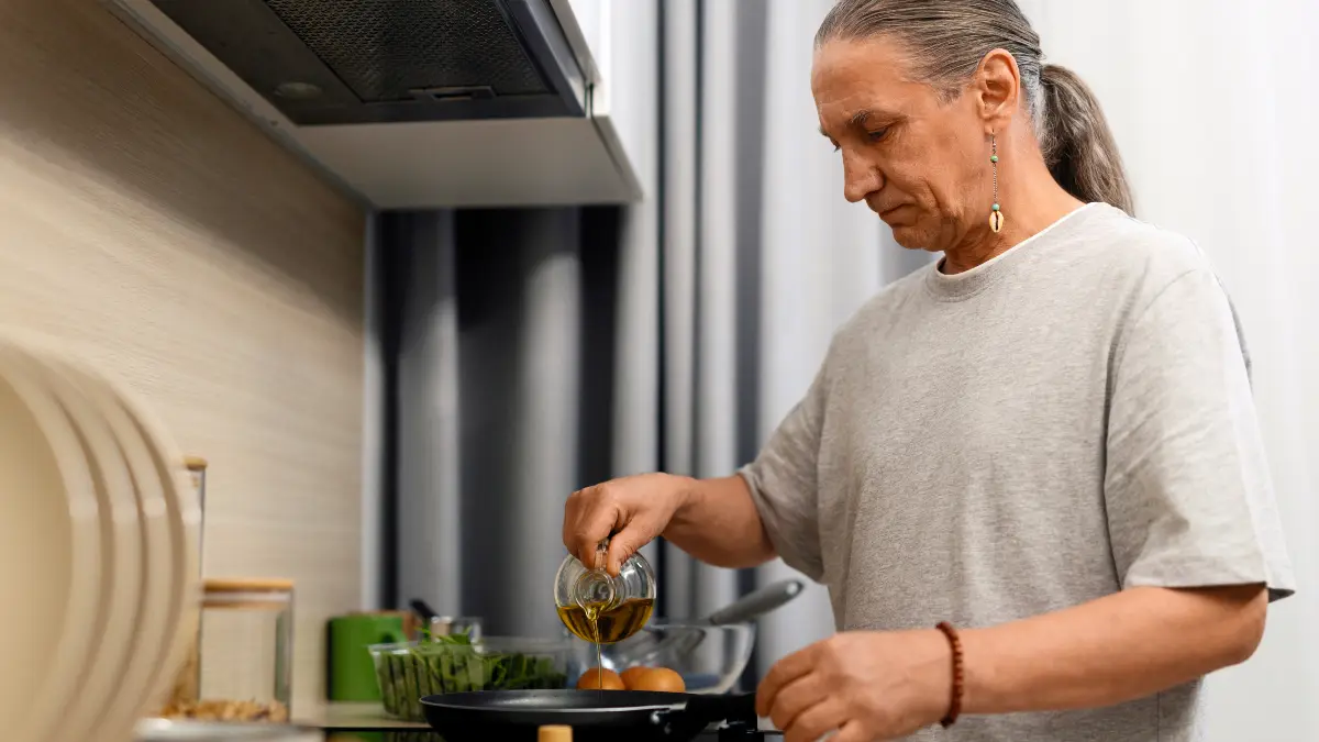 Man in his late 50s pouring golden olive oil from a glass pourer into a pan with eggs on a home stovetop, using extra virgin olive oil for everyday cooking at moderate heat.
