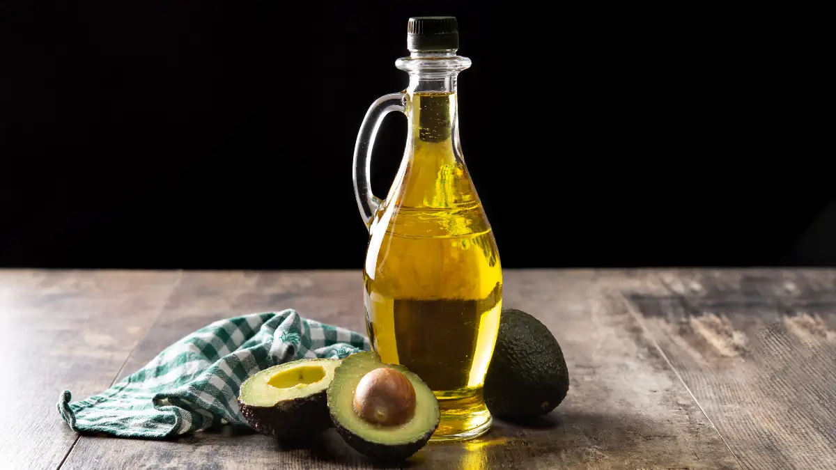 Glass pitcher of golden avocado oil beside a halved avocado on a wooden surface, a stable high-heat cooking oil rich in monounsaturated fat for searing and roasting above 400°F.