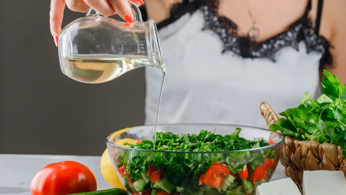 Woman pouring cold-pressed oil from a glass pitcher over a fresh herb salad with tomatoes, cucumber, and parsley, preserving polyphenols by using oil raw as a finishing drizzle.