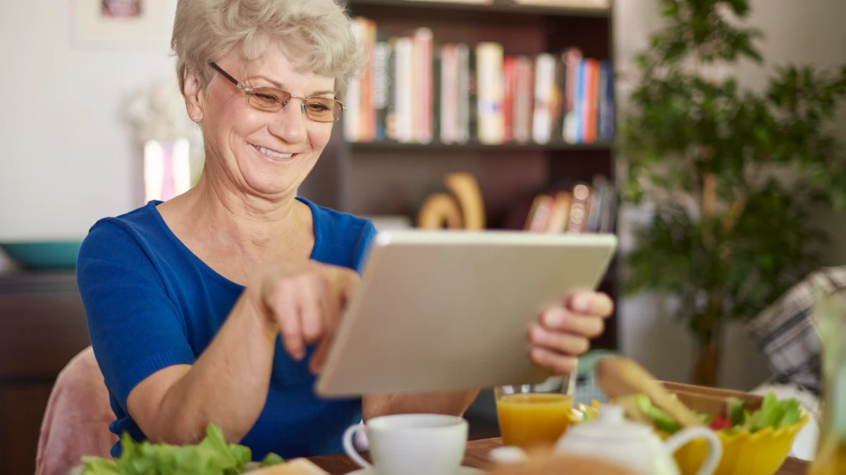 Senior woman using a tablet at the dining table during a healthy meal, demonstrating a scheduled video dinner to lower cortisol.