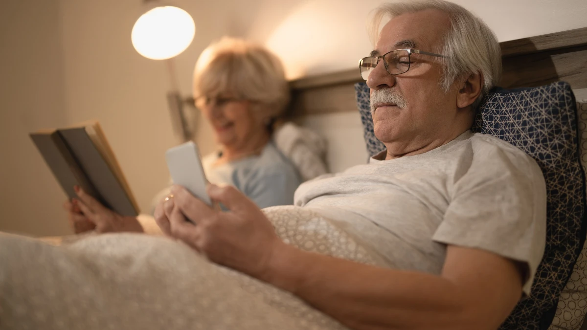 An older man in glasses lies in bed scrolling on his smartphone while his wife reads a book beside him under a warm bedside lamp illustrating how screen use at night can disrupt sleep.