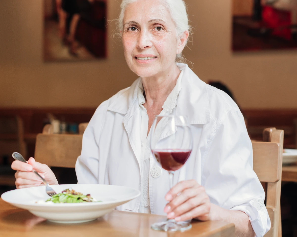 A senior woman dining alone at a wooden table, holding a fork and a glass of red wine while eating a fresh salad to practice mindful, slow eating habits.
