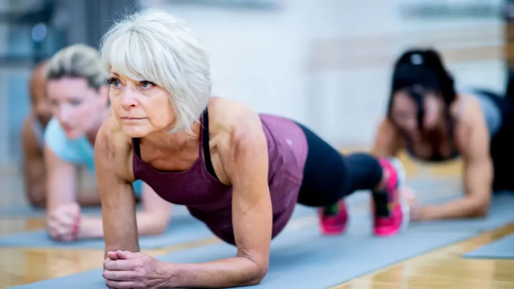 A fit woman in her 50s with short white hair holding a plank position in a group fitness class, illustrating the advice to avoid intense exercise within 3 to 4 hours of bedtime, as hard workouts raise core body temperature and can disrupt sleep.