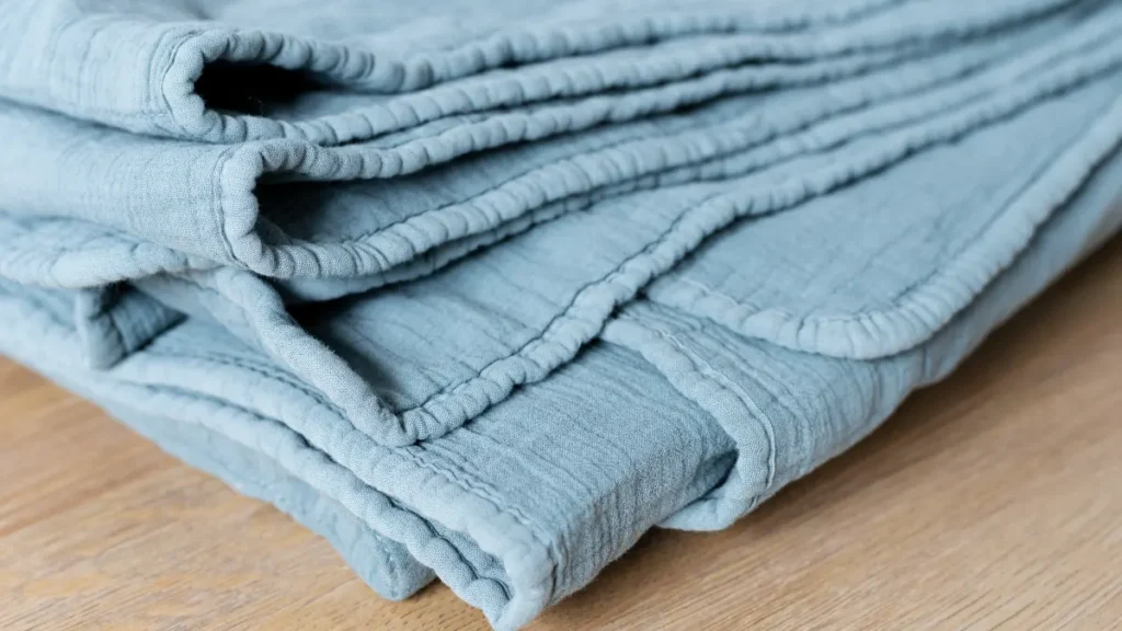 A close-up of a soft, folded light blue cotton muslin blanket resting on a wooden surface, illustrating the recommendation to switch to breathable natural fabric bedding like cotton, bamboo, or linen to stay cooler and reduce heat buildup during sleep.