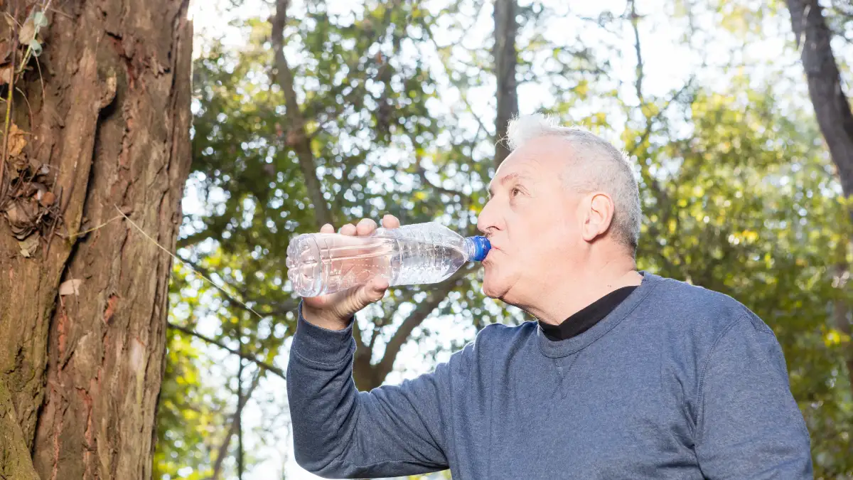 A mature man drinks from a water bottle while outdoors to increase daily hydration and prevent kidney stone growth.