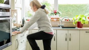 Woman in her late 50s practicing a single-leg stand at her kitchen counter, demonstrating the no-equipment fall prevention balance exercise described in this guide.