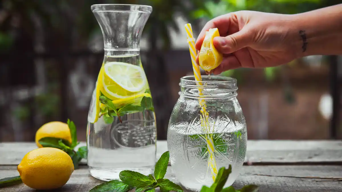 A tattooed hand squeezes a fresh lemon wedge into a glass jar of water to increase citrate levels for optimal kidney health.