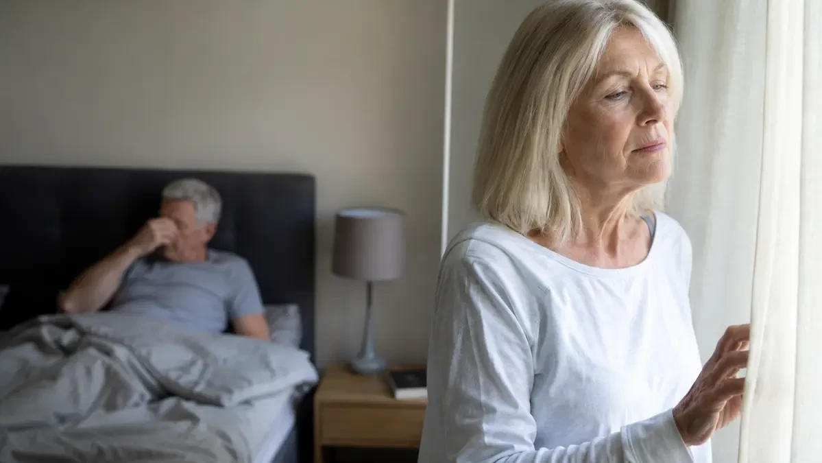 Mature woman looking out a bedroom window in the morning while her husband remains in bed to illustrate the experience of unexplained fatigue after 50 caused by a cellular energy leak.