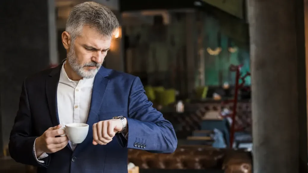 Man over 50 holding a coffee cup and checking his watch in a café, illustrating the importance of caffeine cutoff times for better sleep after 50.
