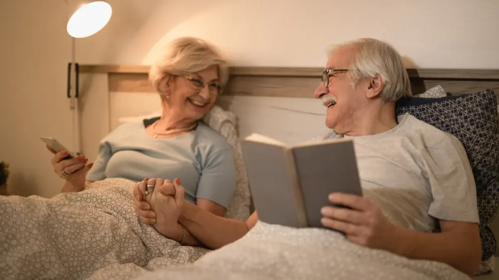Older couple in their late 60s holding hands in bed under warm lamp light, one reading a book and one resting, sharing a bedroom while maintaining their own evening routines.